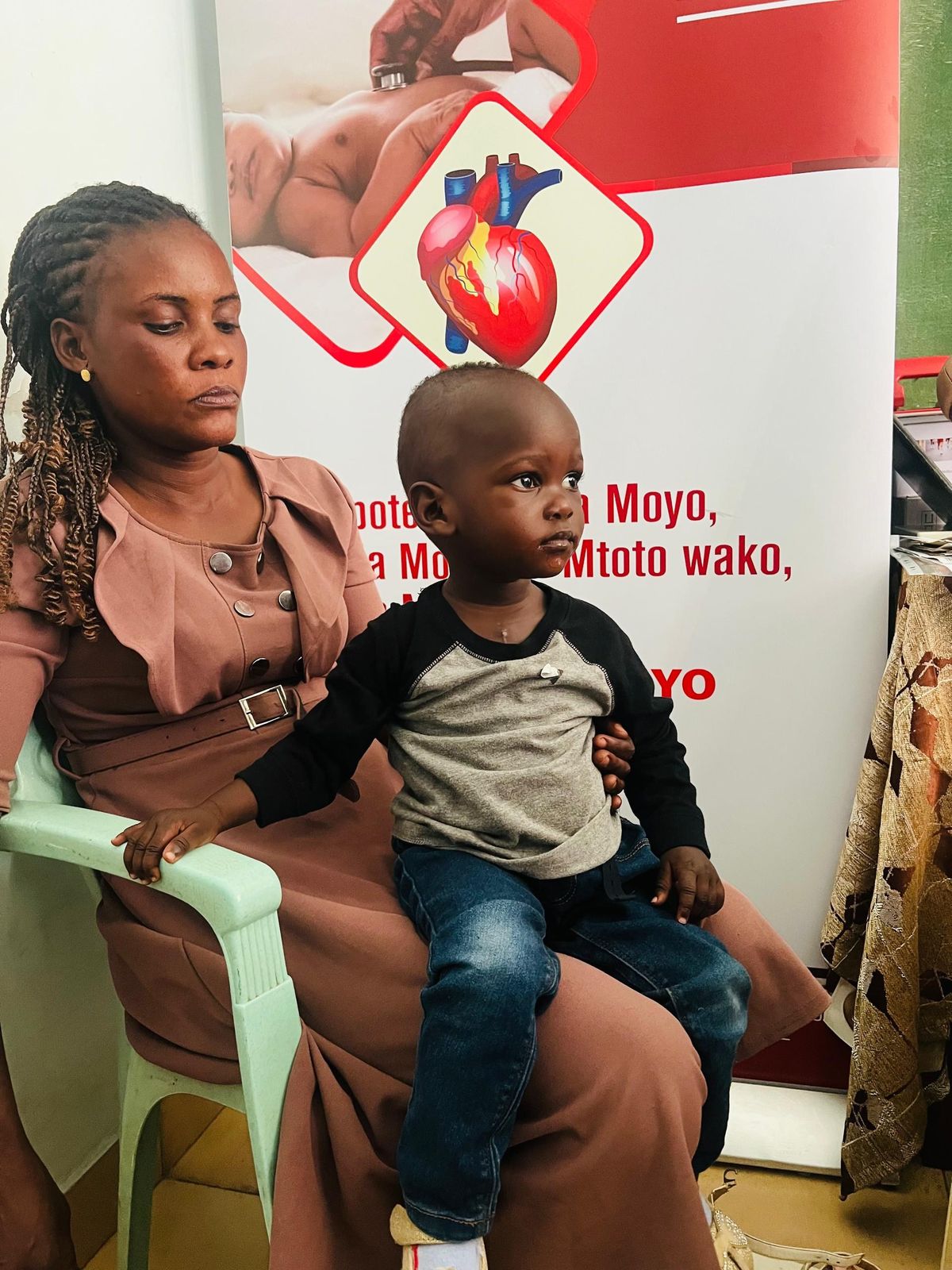 Catherine sits on her mother’s lap beside a One New Heart awareness banner