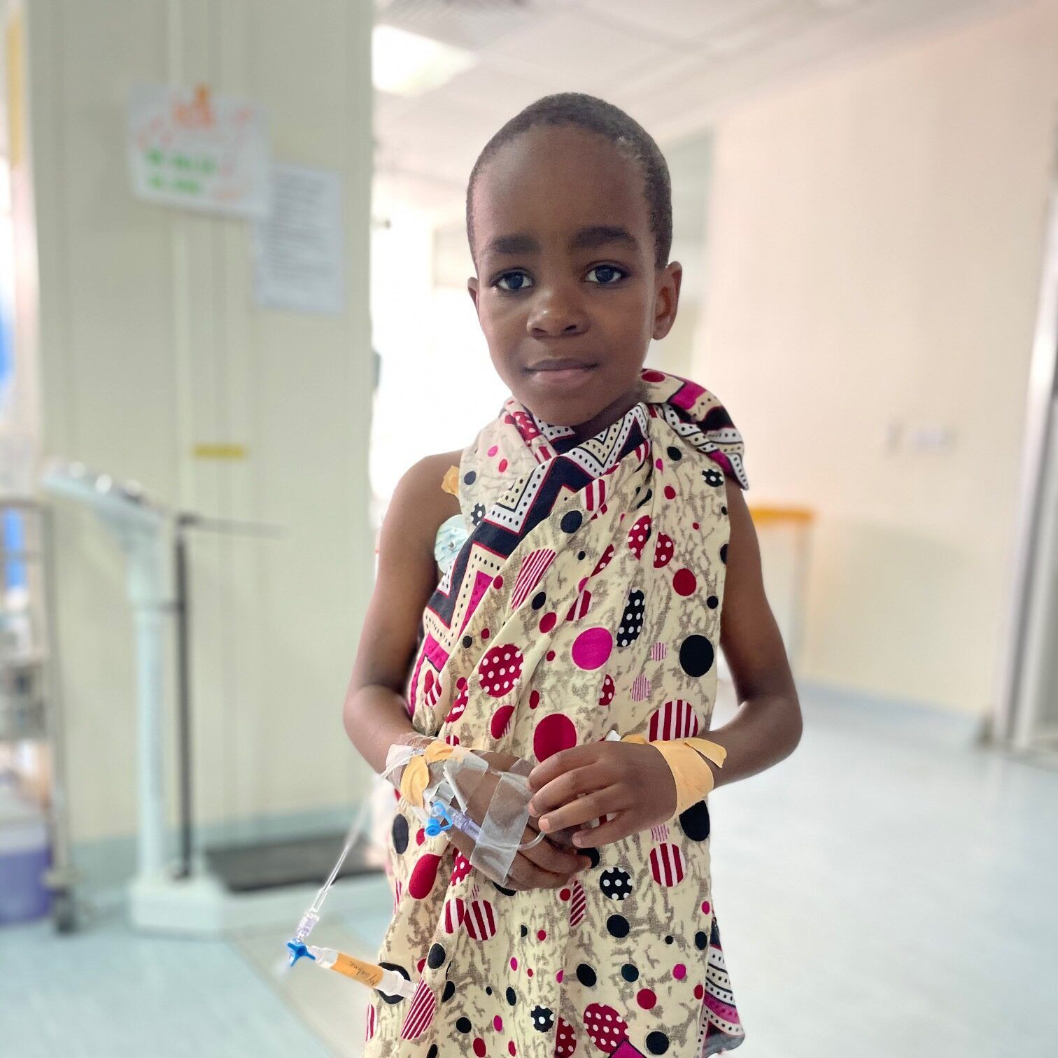 A girl in a patterned dress stands in a hospital corridor, holding a cup